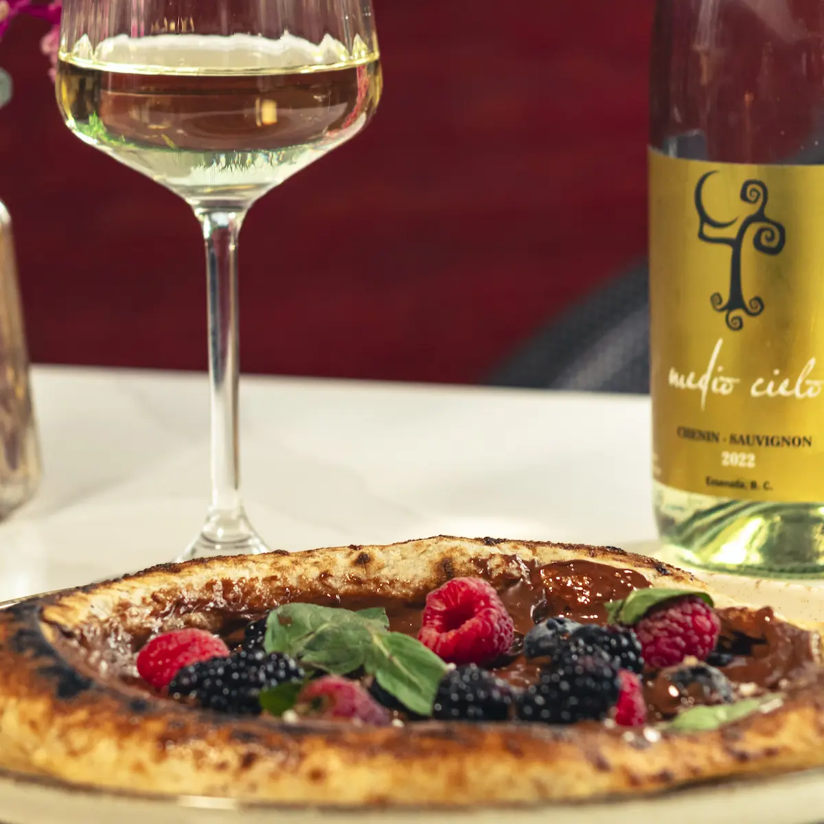 Copa de vino blanco y botella junto a una mini pizza dulce con frutos rojos.