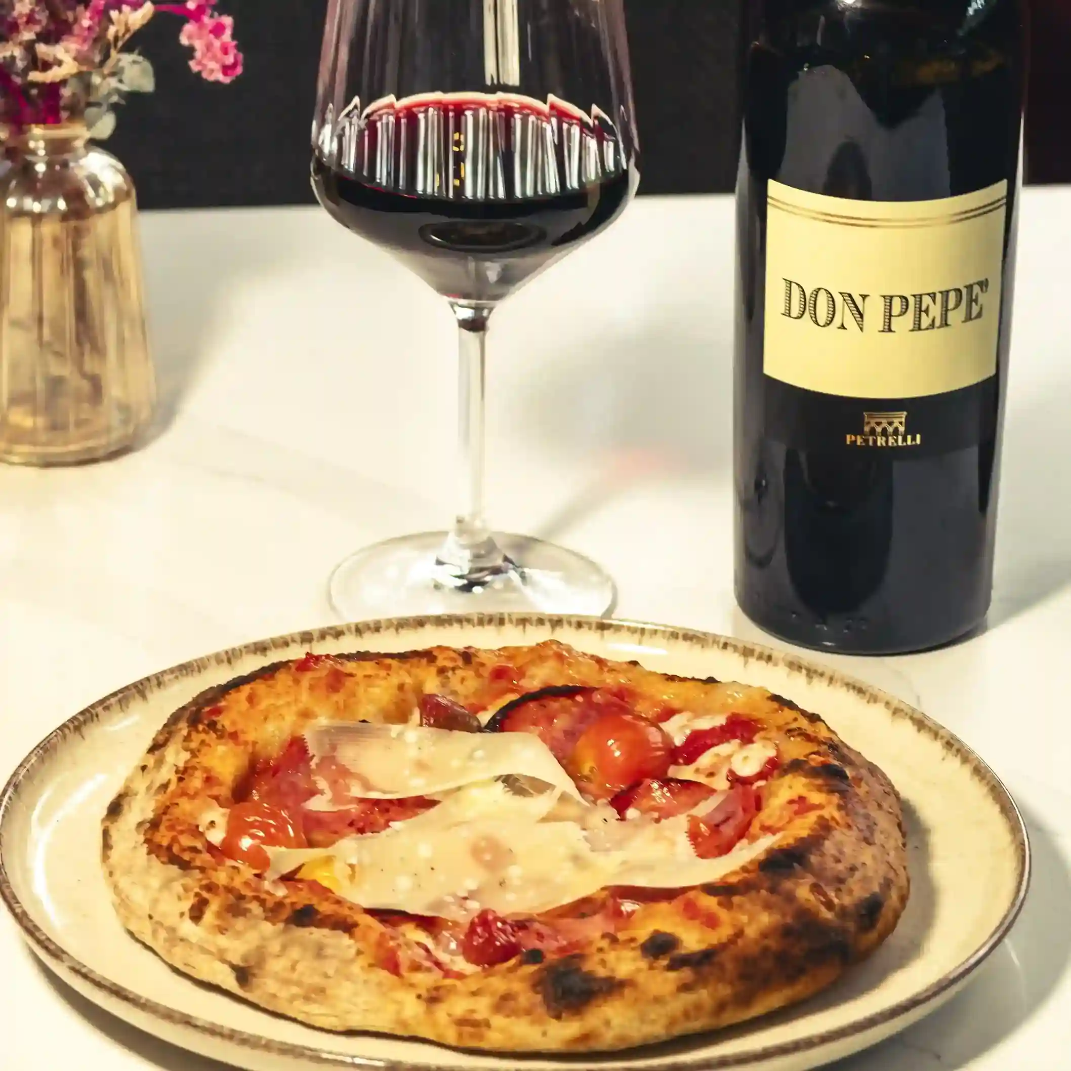 Copa de vino tinto y botella Don Pepe junto a una mini pizza salada con jamón y queso.