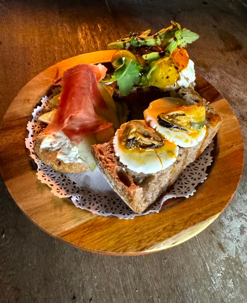 Tabla de madera con pan rústico, queso de cabra, jamón serrano y ensalada de hojas verdes.