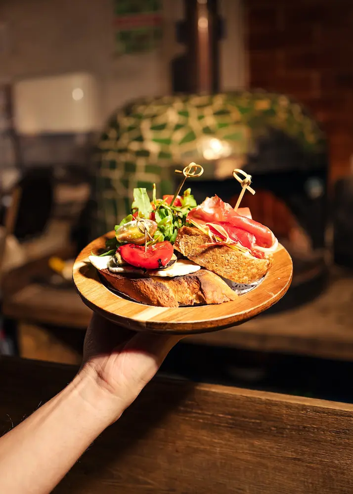 Una tosta con jamón serrano y una tosta con pimiento asado y hojas verdes, servidas en un plato de madera.