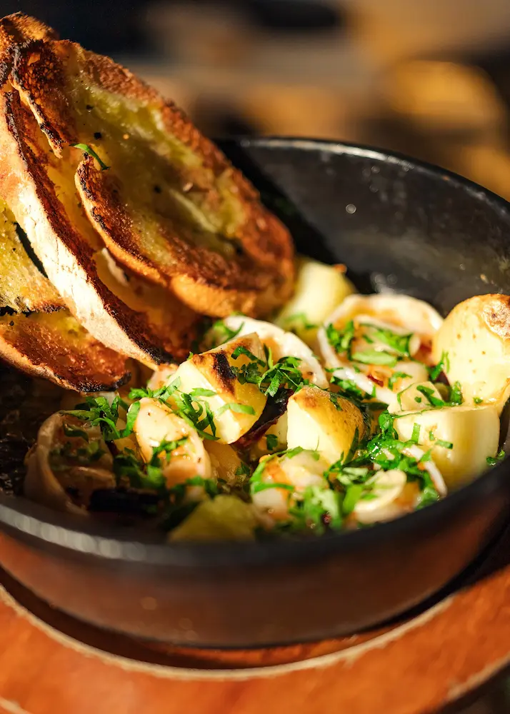 Plato de camarones y papas en una salsa con hierbas, servido en un tazón de hierro fundido con rebanadas de pan tostado.