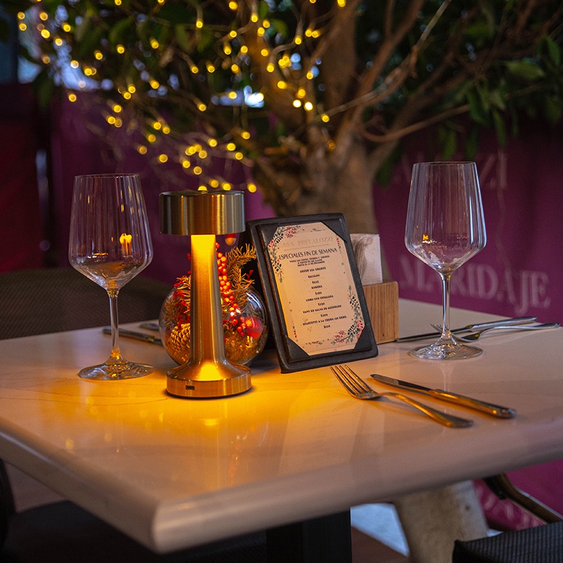 Mesa elegante con copas de vino, cubiertos dorados y menú iluminado por una lámpara dorada, con luces cálidas y árbol de fondo.