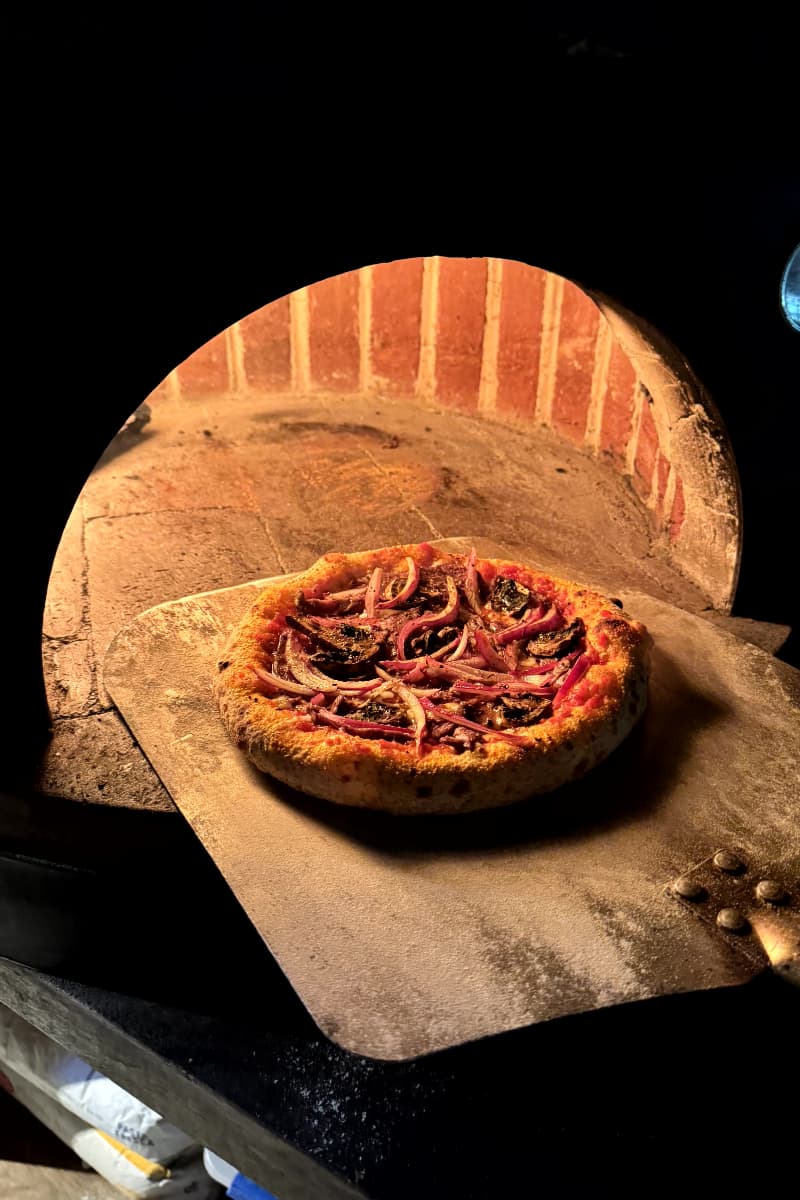 Pizza artesanal horneándose en horno de leña con cúpula de ladrillo, colocada sobre una pala de madera.
