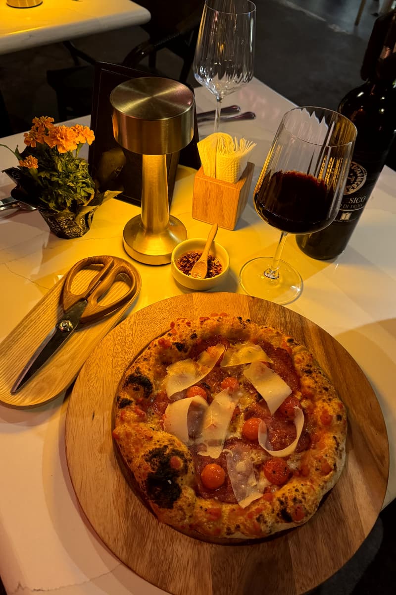 Pizza con tomates, champiñones y queso sobre una mesa con una copa de vino tinto y condimentos.