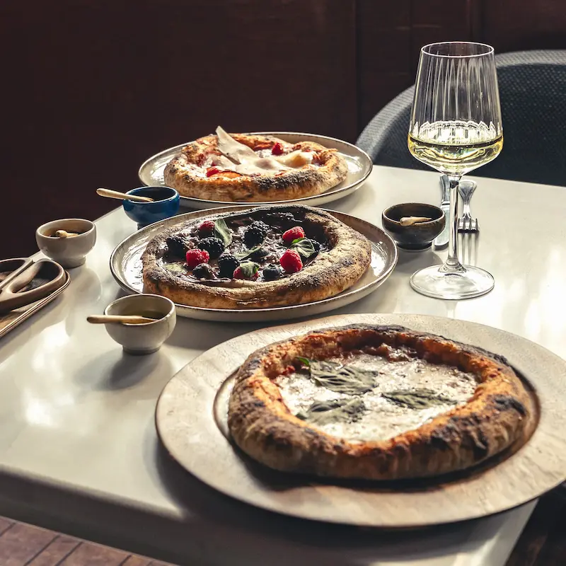 Mesa servida con varias pizzas artesanales recién horneadas, acompañadas de vino blanco y un ambiente acogedor.