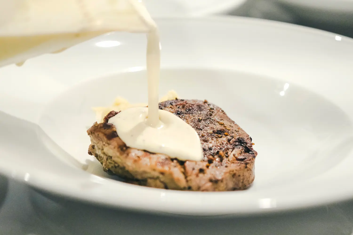 Salsa cremosa siendo vertida sobre un plato de carne a la parrilla servido en un plato blanco.