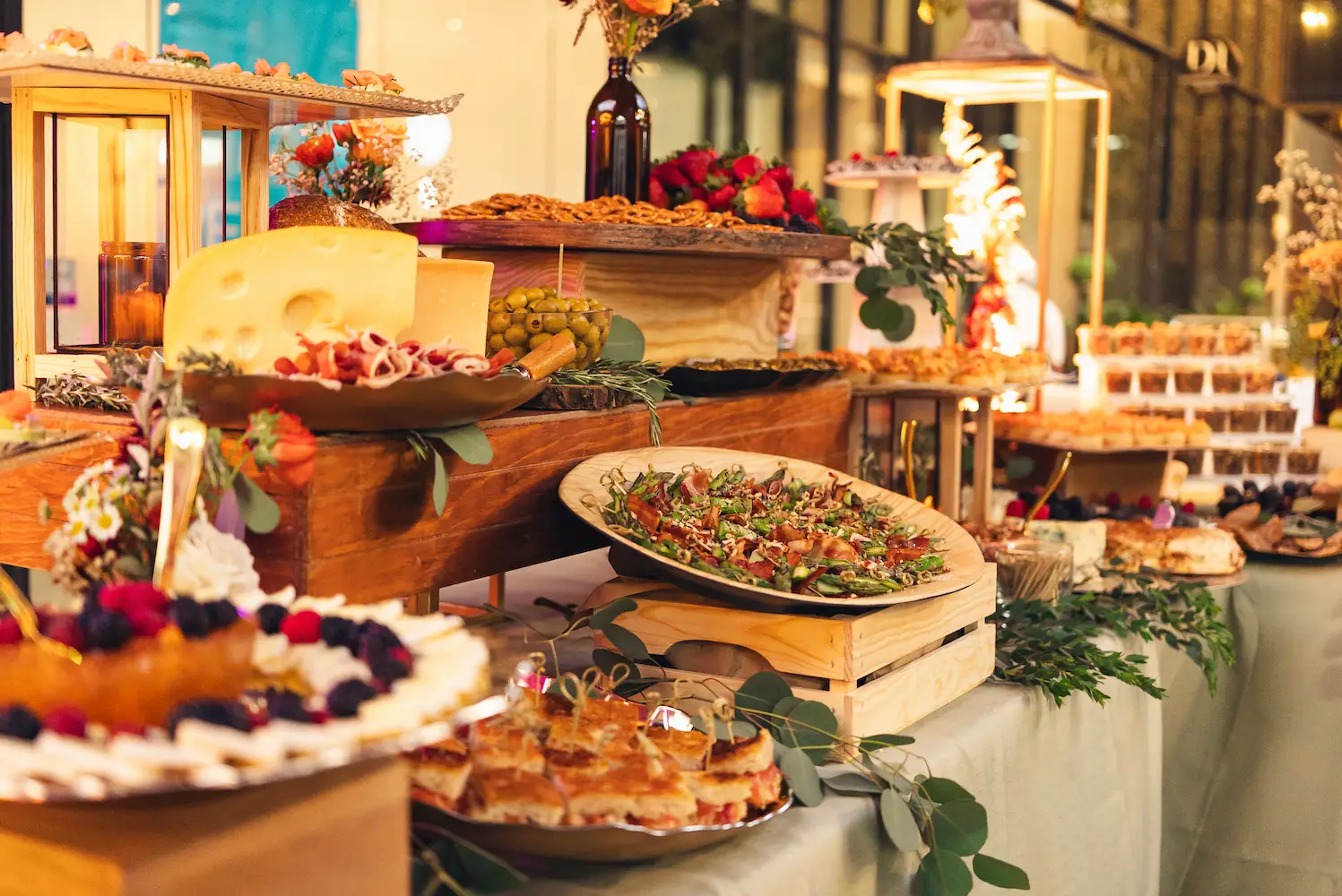 Mesa de buffet festivo en un evento nocturno con varias fuentes de comida, incluyendo tartas y aperitivos, decorada con un ambiente cálido.